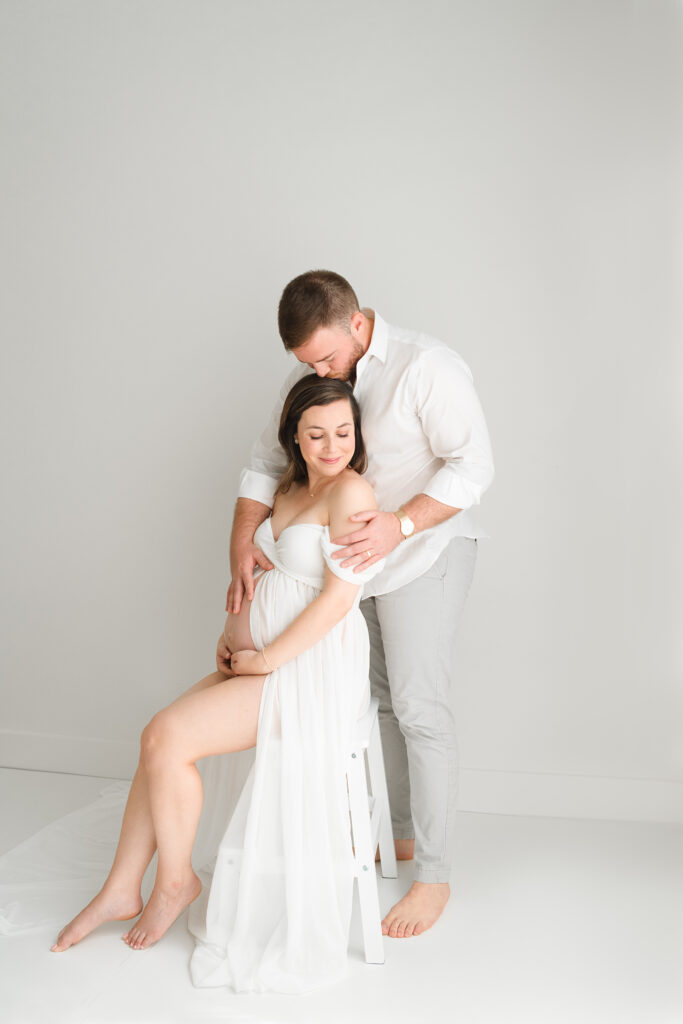 Pregnant mother wearing a white dress posing for a photograph with her husband in a Jacksonville photography studio