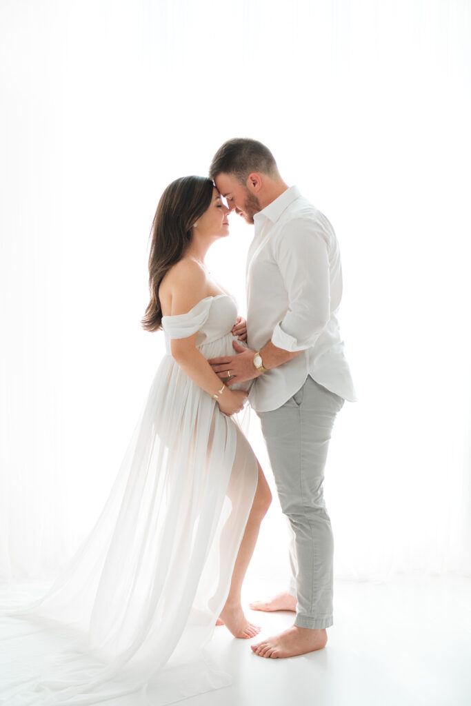 Pregnant mother wearing a white dress posing for a photograph with her husband in a Jacksonville photography studio